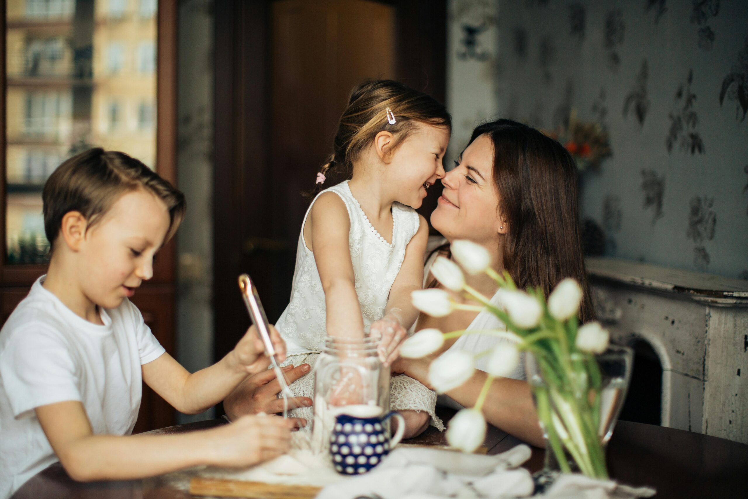 A mother smiles as she shares a tender moment with her young daughter, while another child sits at the table drawing.
